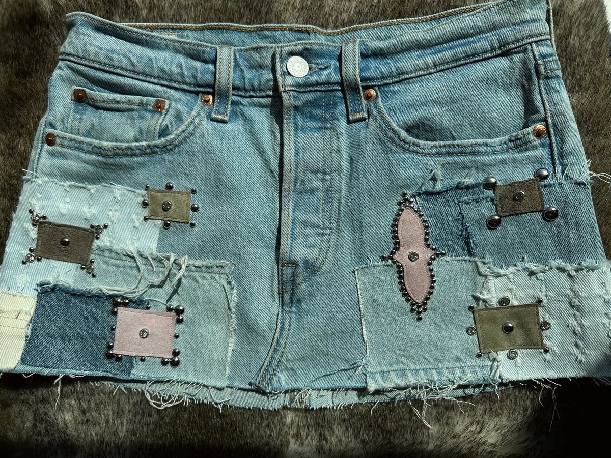 Full view of a reworked denim mini skirt with frayed edges, pink and neutral patchwork, and metal hardware.